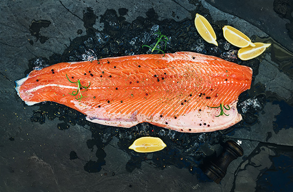 Bright pink salmon surrounded by lemons and sprinkled with spices on a black slab.