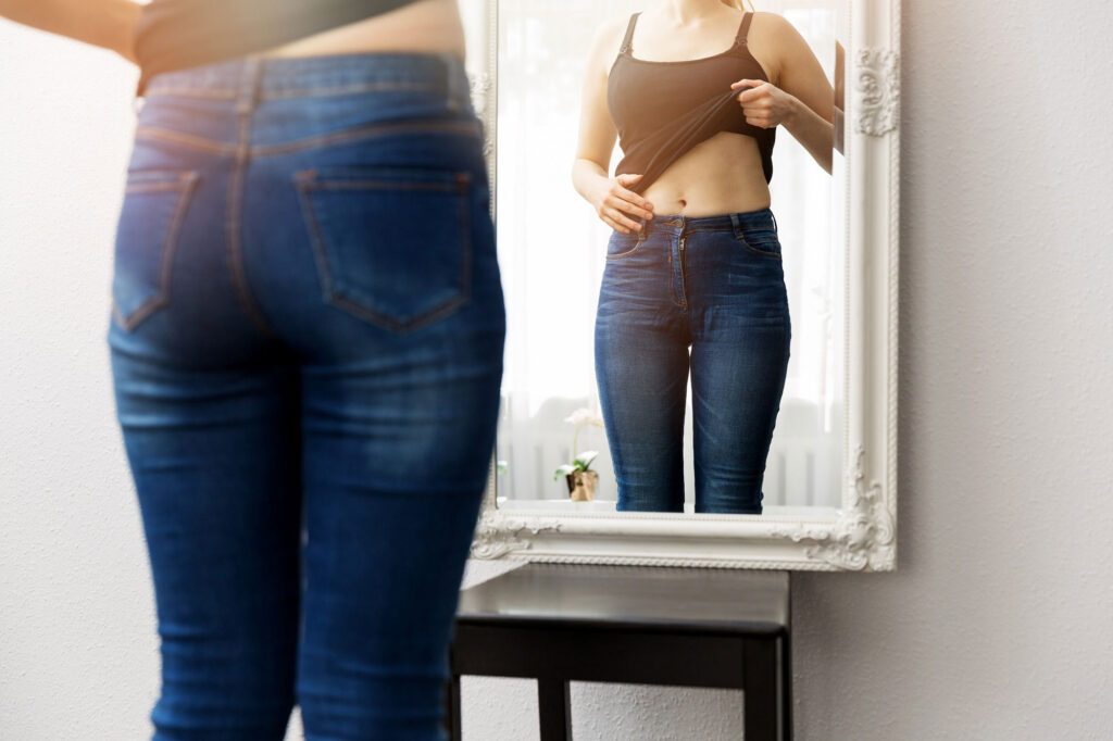 Woman looking in mirror while holding up shirt to reveal body after weight loss surgery.