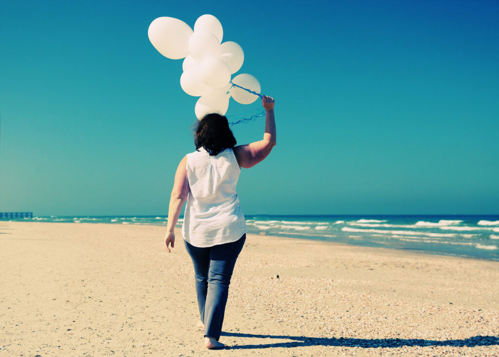 Woman walking on beach holding white balloons while reflecting on life after weight loss surgery.