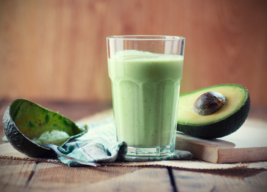 Avocado smoothie drink between an empty avocado skin and an avocado half on a cutting board.