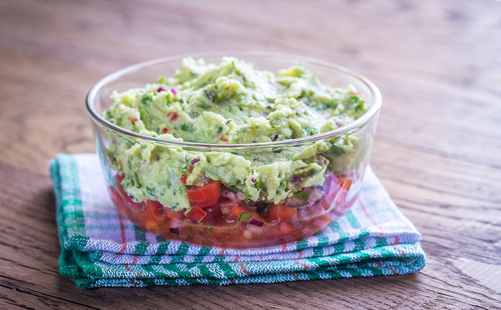 Avocado spread over tomatoes in a glass bowl sitting on a dish rag.