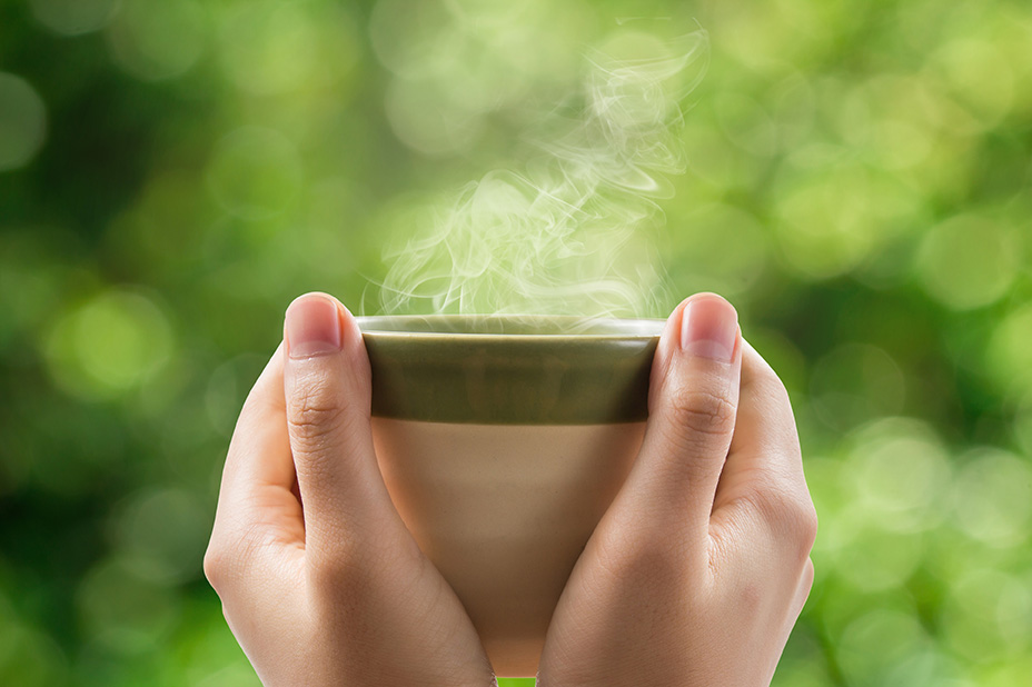 Close up of hands holding a green and beige cup of tea with smoke coming out of it.