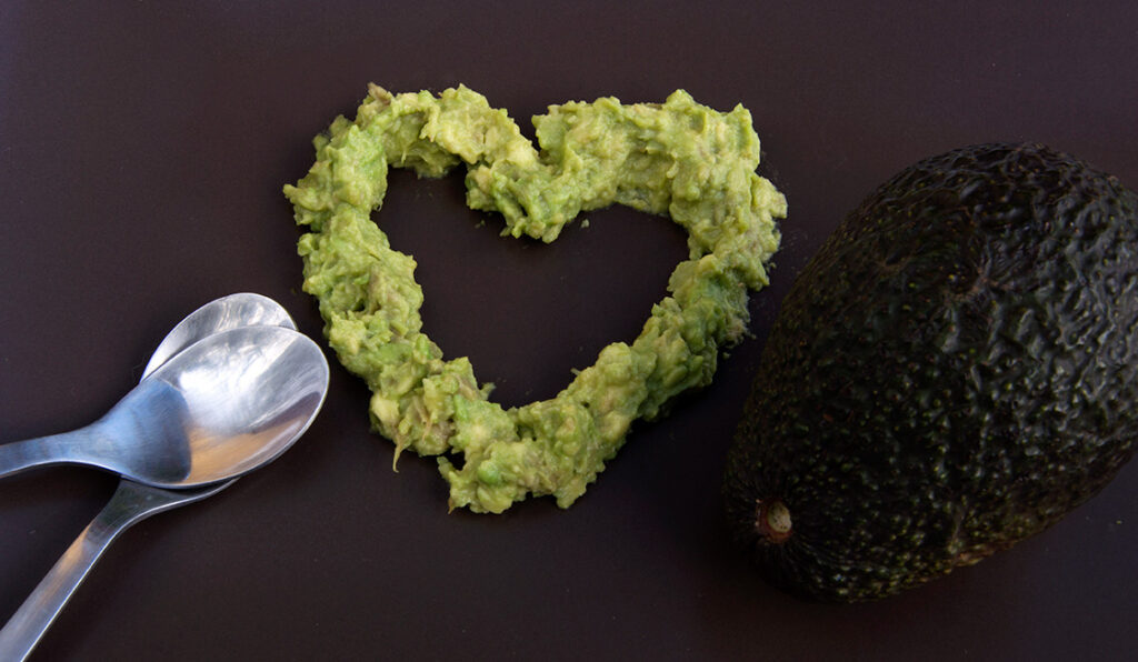 Avocado mashed up in the shape of a heart in between two silver spoons and an avocado.
