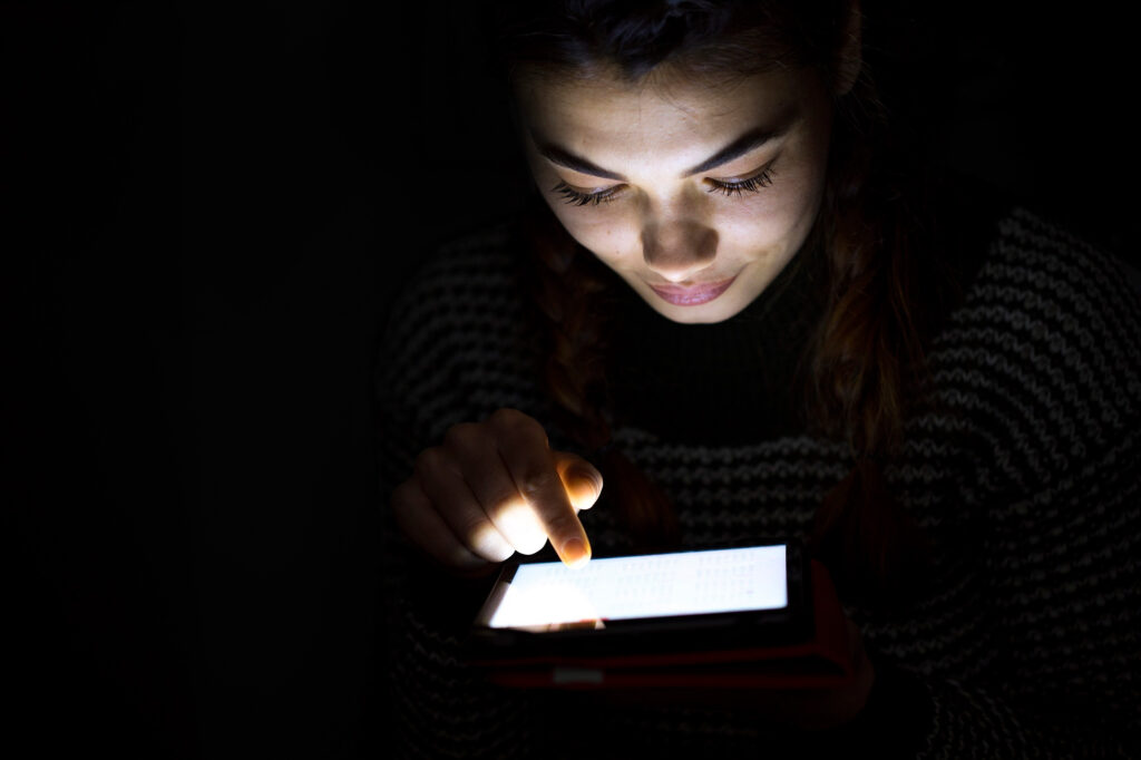 Woman sitting in the dark while looking at a lit up tablet.