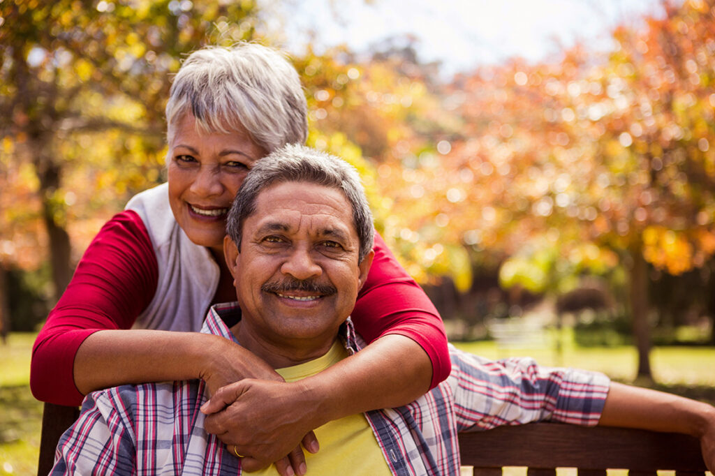 Senior Hispanic woman hugging around the neck of husband sitting on bench.