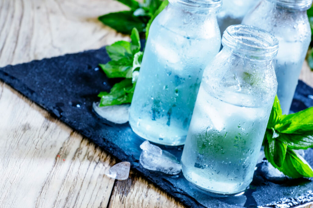 Three cold glasses of water surrounded by ice and leaves sitting on a blue cloth.