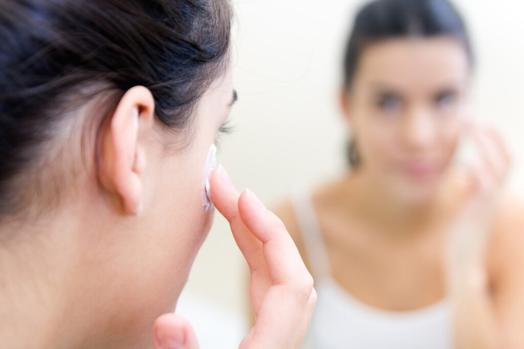 Woman looking in mirror and putting cream under her eyes to follow summer skin care tips.