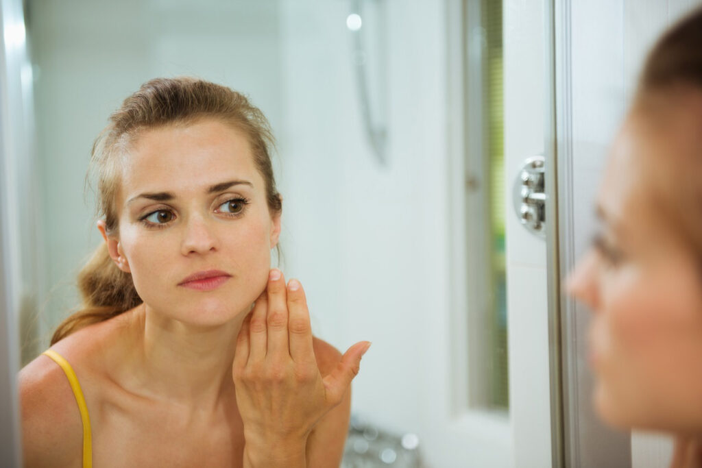 Woman looking in mirror and examining face to follow summer skin care tips.
