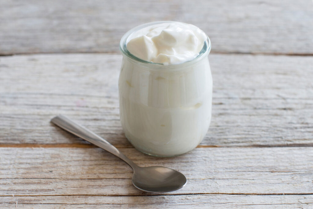 Jar of plain Greek yogurt and silver spoon sitting on a wooden table.