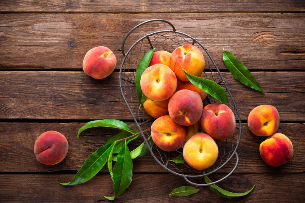 Basket of peaches with a few other peaches and leaves sitting on a wooden table.