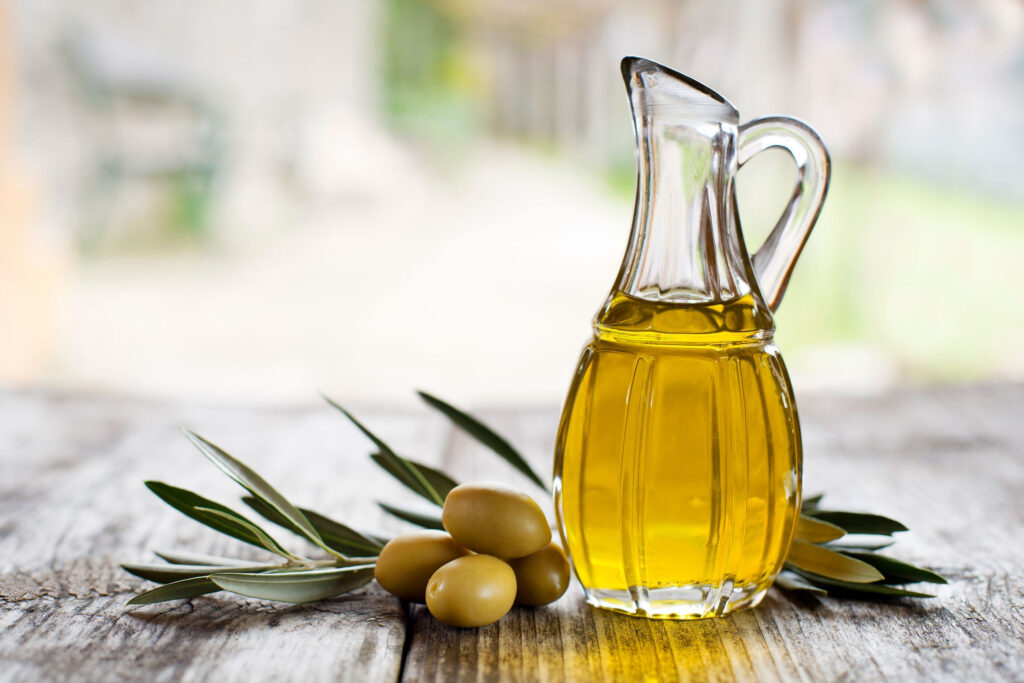 Pitcher of olive oil surrounded by olives on a wooden table.