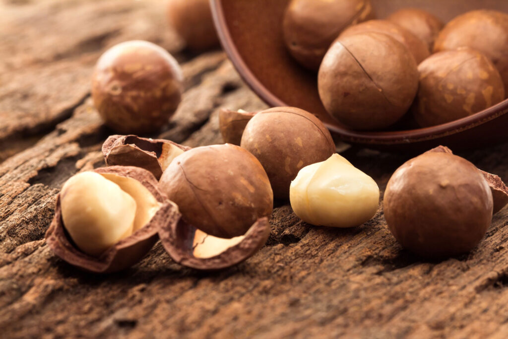 Broken macadamia nuts roll out of brown bowl onto wood.