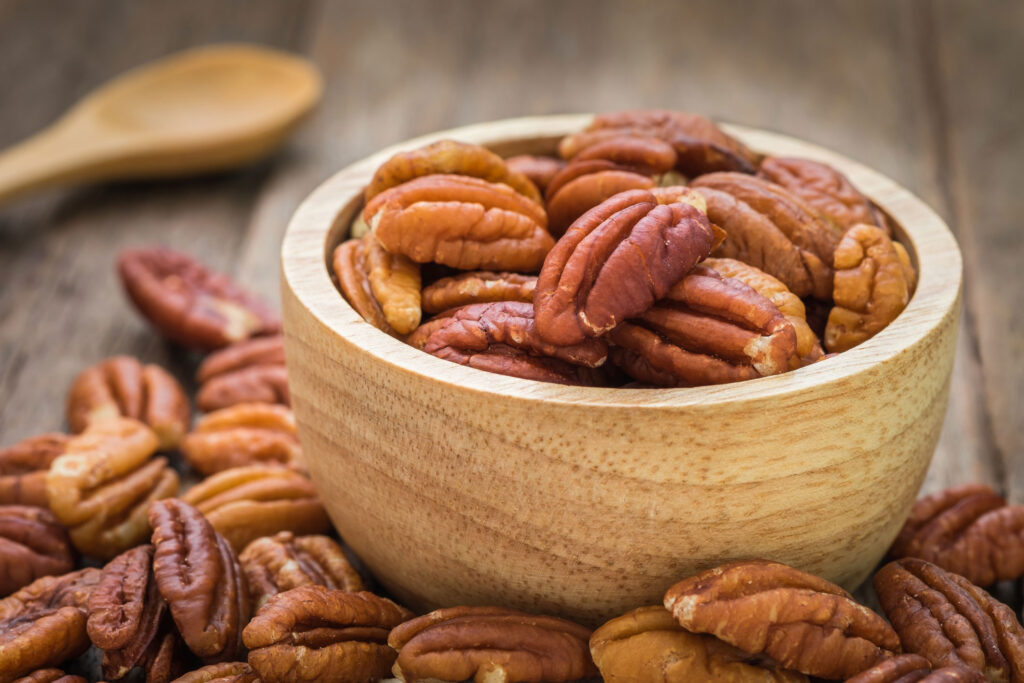 Pecans surround a light colored wooden bowl filled with more pecans.