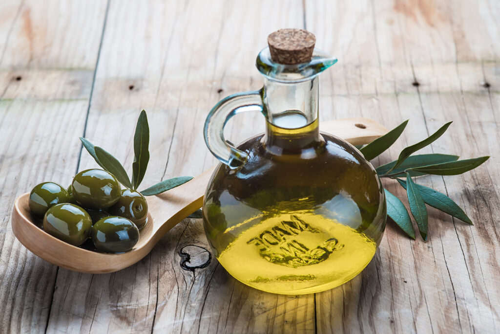 Jar of olive oil and a spoon with olives sitting on a wooden table.