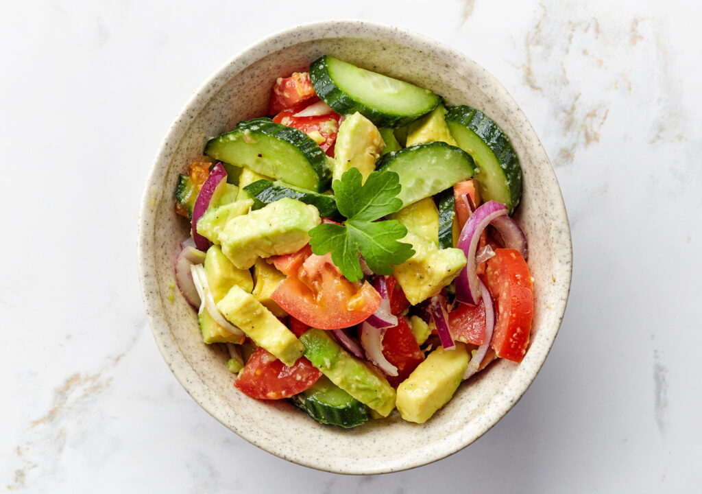 A bowl of fresh avocados and vegetables sitting on a marble countertop.