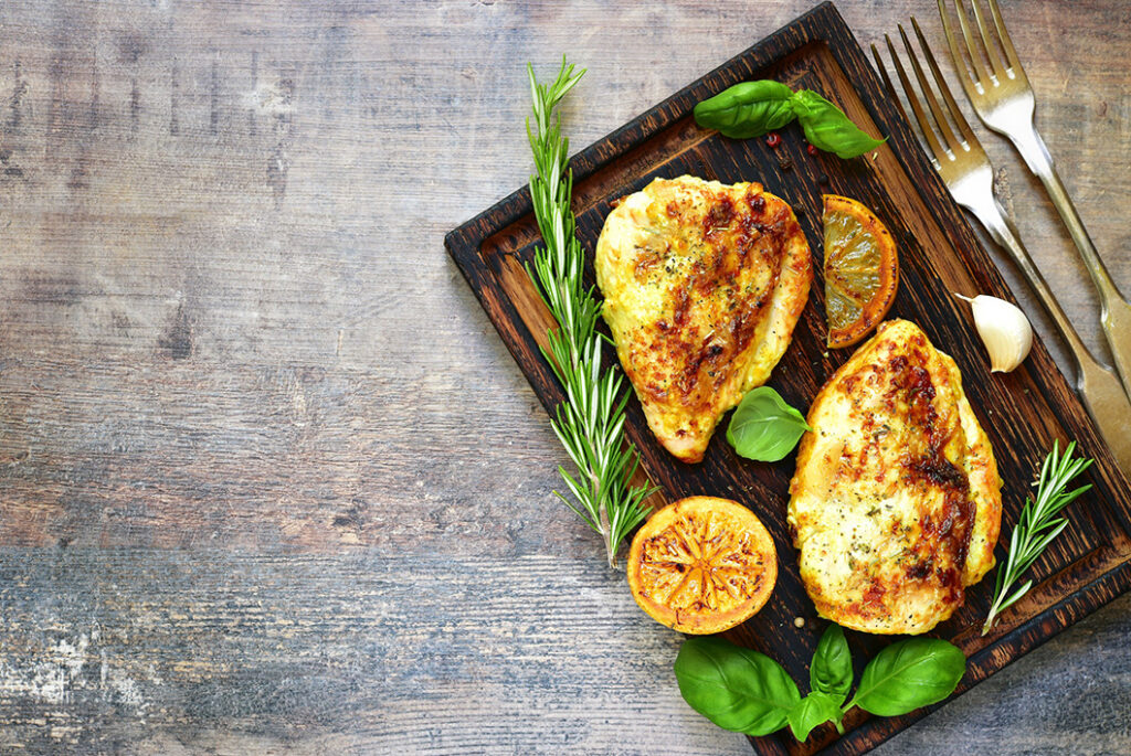 Cutting board with grilled chicken surrounded by rosemary and lemons.