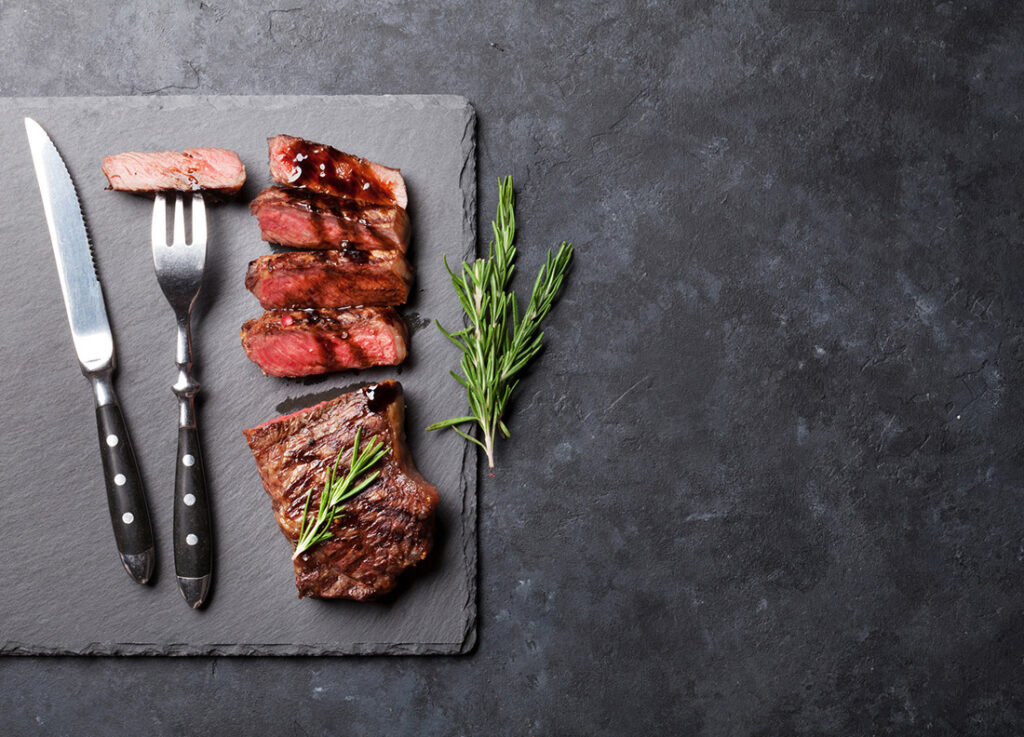 Sirloin cut up next to fork and knife on concrete slab.