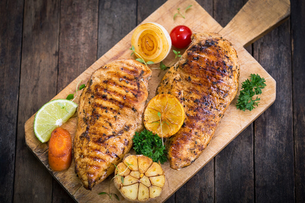 Two pieces of grilled chicken surrounded by lemons and vegetables on cutting board.