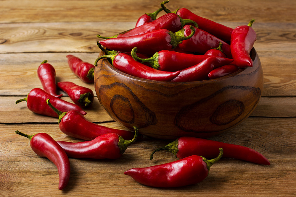 Wooden bowl of red chili peppers with some scattered chili peppers around the bowl on a wooden table.